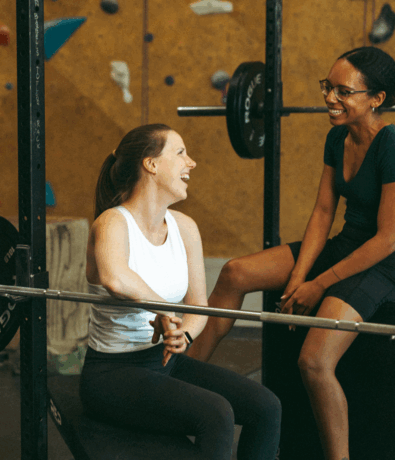 Two women at a gym are smiling and chatting. One sits on a bench next to a barbell, while the other sits on a nearby platform. Climbing holds are visible behind them as they discuss their new fall membership.