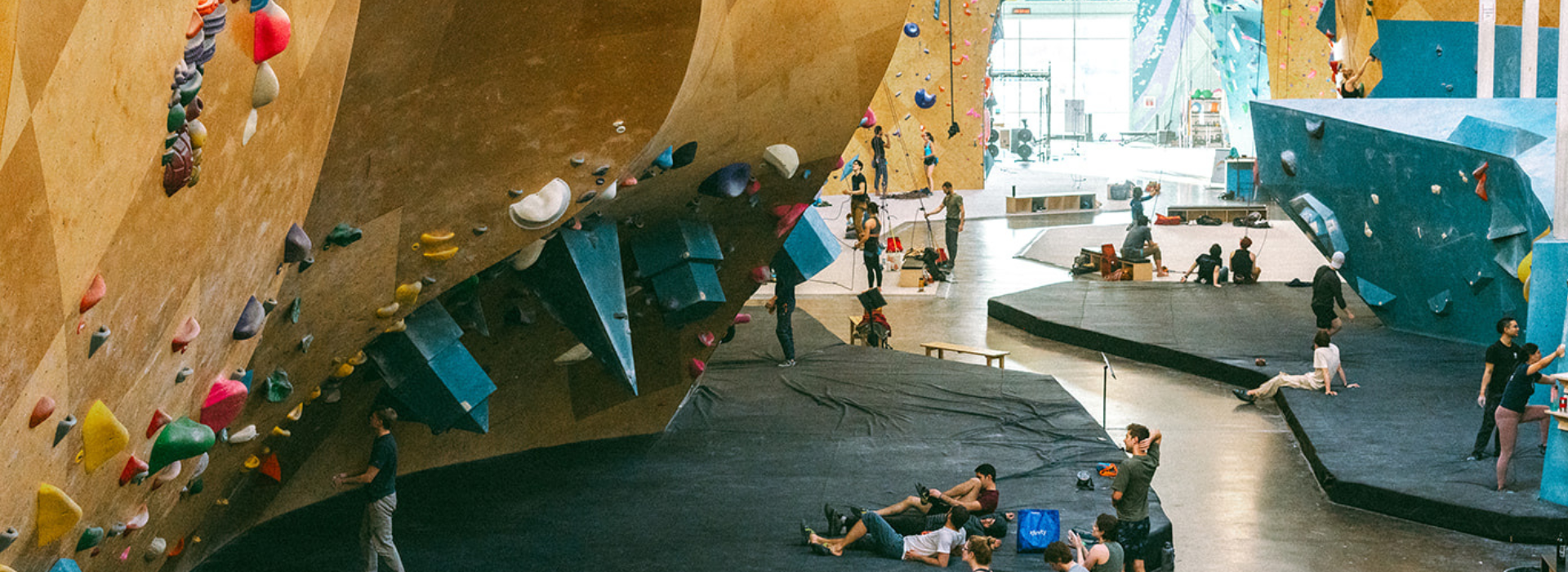 People are climbing and relaxing inside a large indoor rock climbing gym with colorful holds, high walls, and padded flooring. Some climbers are on the walls while others sit or stand on the mats below, making the most of their fall membership.