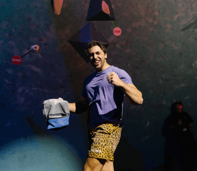 A man in a purple shirt and leopard-print shorts smiles and gestures triumphantly with a chalk bag at Boulderfest 2025 in Minneapolis, standing before an indoor climbing wall with holds labeled START and ZONE. Another person appears in the background.