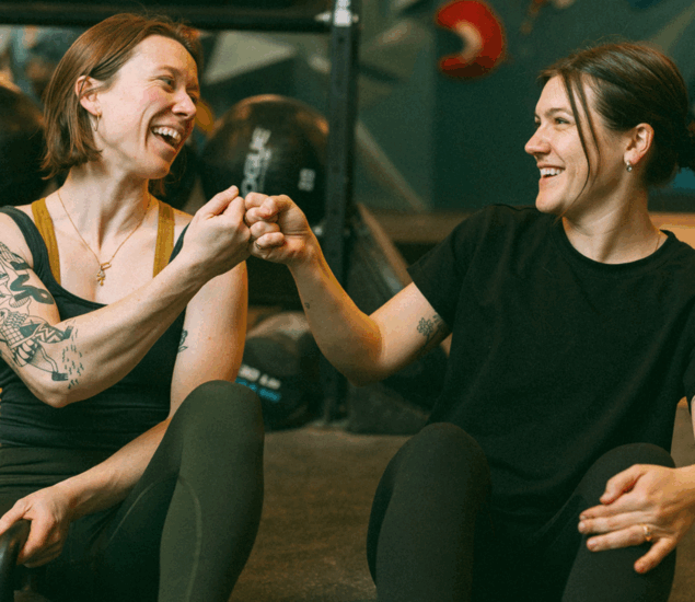 Two women in athletic wear sit on a gym floor in Brooklyn, smiling and fist-bumping each other. Both have tattoos on their arms and appear happy and relaxed, suggesting camaraderie after private instruction or a workout.