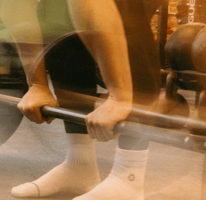 A person in white socks grips a barbell, preparing to lift in a Brooklyn gym. The image’s motion blur effect highlights the energy of private instruction and focused movement.