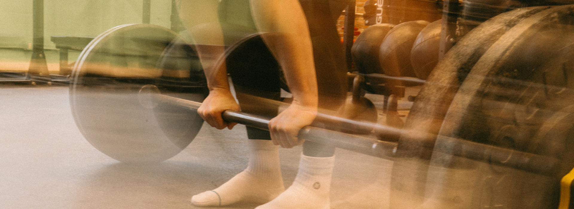 A person in white socks grips a barbell, preparing to lift in a Brooklyn gym. The image’s motion blur effect highlights the energy of private instruction and focused movement.