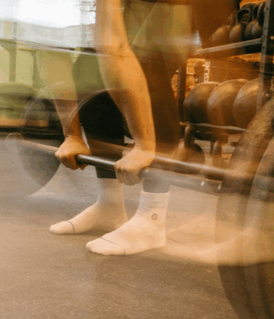 A person in white socks and black leggings lifts a barbell in a Brooklyn gym. The motion blur highlights the intensity of weightlifting, with kettlebells and hints of private instruction visible in the background.
