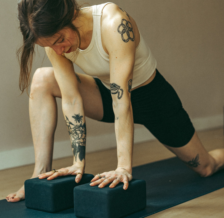 In a sunlit Brooklyn studio, a tattooed individual practices yoga indoors, performing a low lunge with two black yoga blocks on a mat—an ideal setting for private instruction in comfortable attire.