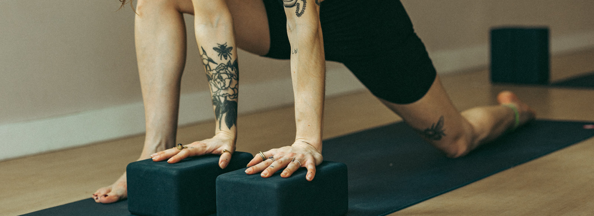 A person with tattoos on their arms and leg practices yoga on a mat in Brooklyn, using two yoga blocks for support while performing a low lunge stretch, suggesting the focus and care of private instruction indoors.