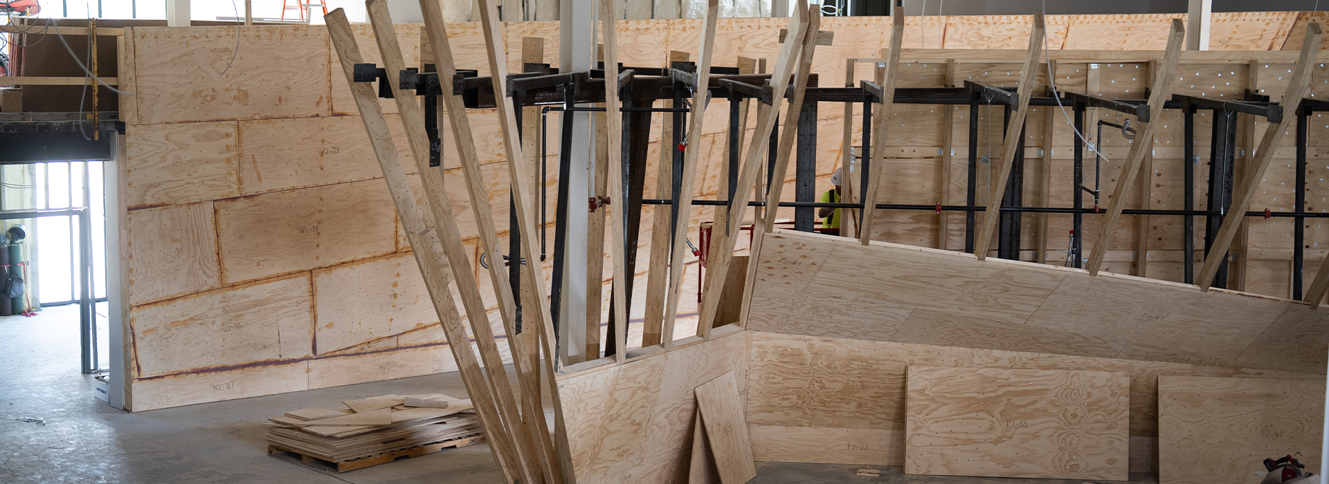 Stacks and sheets of plywood are arranged to form large, angular wooden structures inside an unfinished indoor space in St. Paul, suggesting construction or set building in progress.