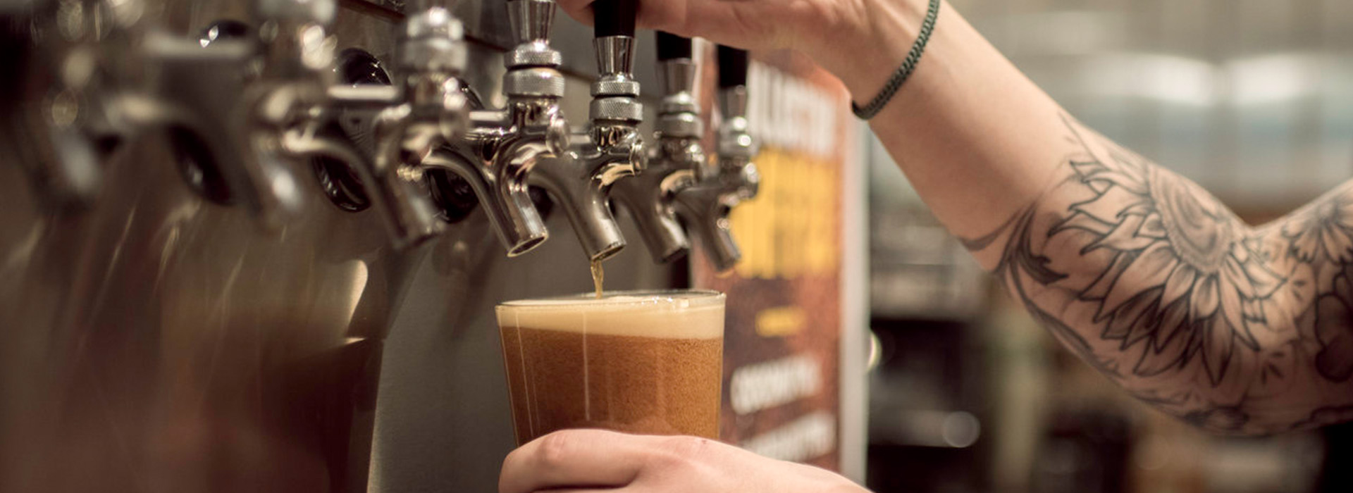 A tattooed person’s hand holds a plastic cup under a row of beer taps at West Wall Cafe, filling it with foaming draft beer. The blurred background hints at the lively ambiance of this popular cafe bar.