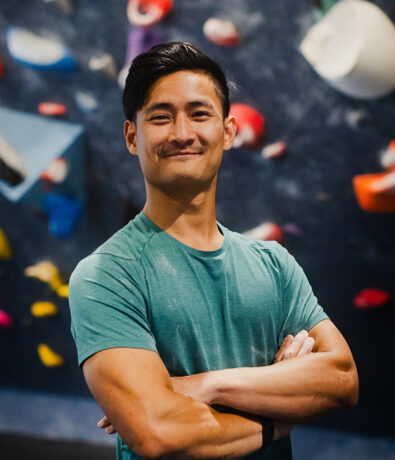 A young man with short dark hair stands confidently with arms crossed, wearing a teal t-shirt. The background shows a colorful indoor climbing wall in Seattle, perfect for those seeking personal coaching and new challenges.