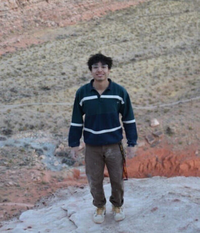 A person with dark hair, wearing a striped sweater, brown pants, and sneakers, stands on a rocky cliff overlooking a desert landscape and hills near Tempe—an inspiring scene for personal coaching and self-reflection.