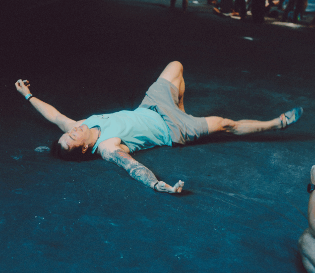 A person in a blue tank top and shorts lies sprawled on their back on a dark floor, arms and legs spread out. Under bright light in an otherwise dim area, they appear relaxed—perhaps unwinding after Boulderfest 2025 in Austin.