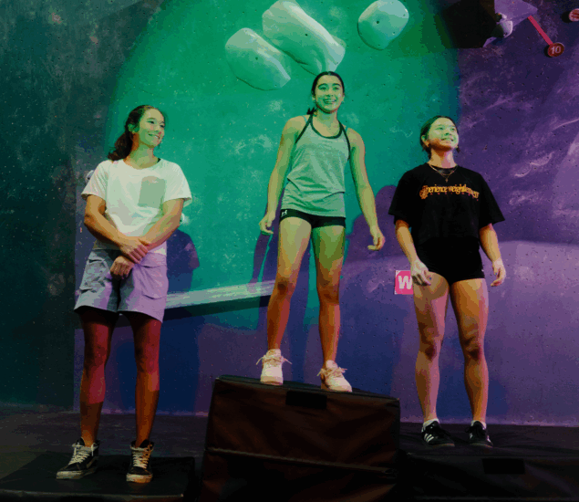 Three young women stand on a podium at an indoor climbing gym in Austin, smiling under green lighting and climbing holds—a perfect Boulderfest 2025 recap, with the center woman standing higher to suggest her first place win.