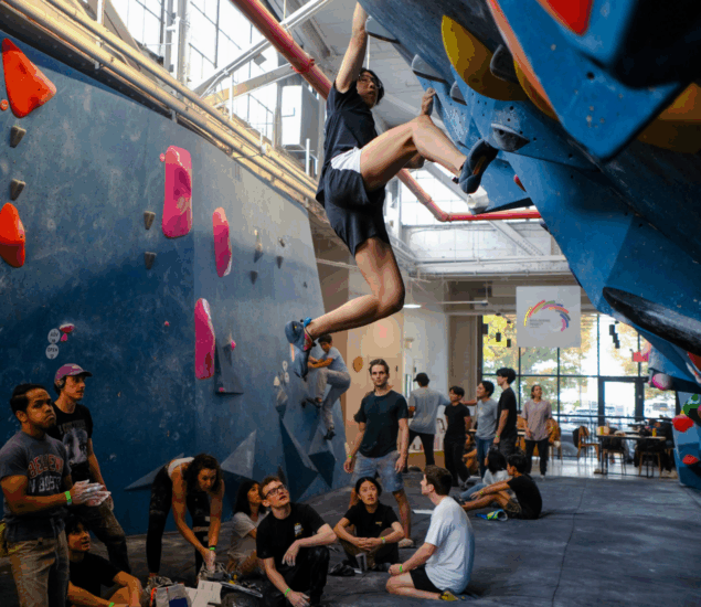 A person climbs a blue indoor bouldering wall at Boulderfest 2025 in Brooklyn as others watch, sit, or prepare nearby. The gym is bustling with people, climbing holds are visible, and sunlight streams through large windows in the background.