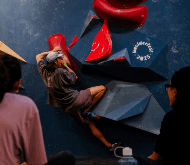 A person wearing a cap and shorts climbs an indoor bouldering wall with red and black holds at Boulderfest 2025 in Brooklyn, watched by two seated people. The event name is displayed on the wall.