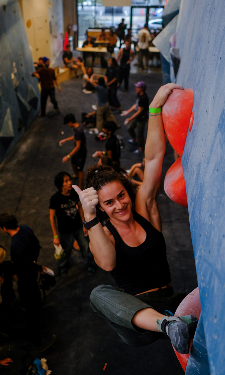 A woman climbs a bouldering wall indoors in Brooklyn, gripping large red holds. She smiles at the camera and gives a thumbs-up. Several people are climbing or watching in the background of the busy gym.