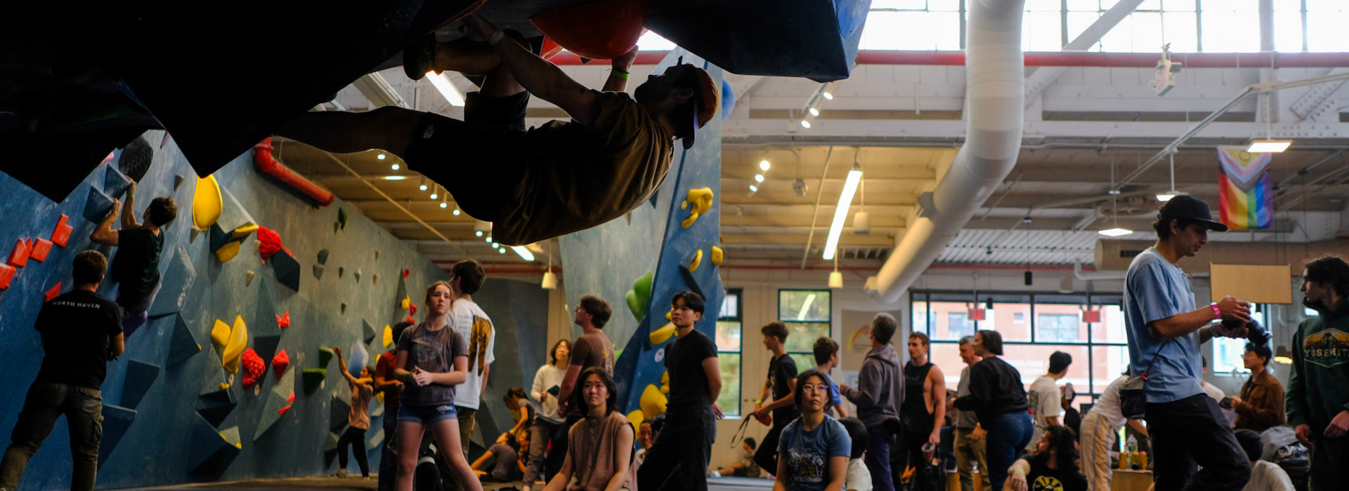 A crowded indoor rock climbing gym in Brooklyn buzzes with energy as climbers tackle colorful walls and others socialize. One climber hangs from an overhang in the foreground—a lively scene fitting for a Boulderfest 2025 recap.