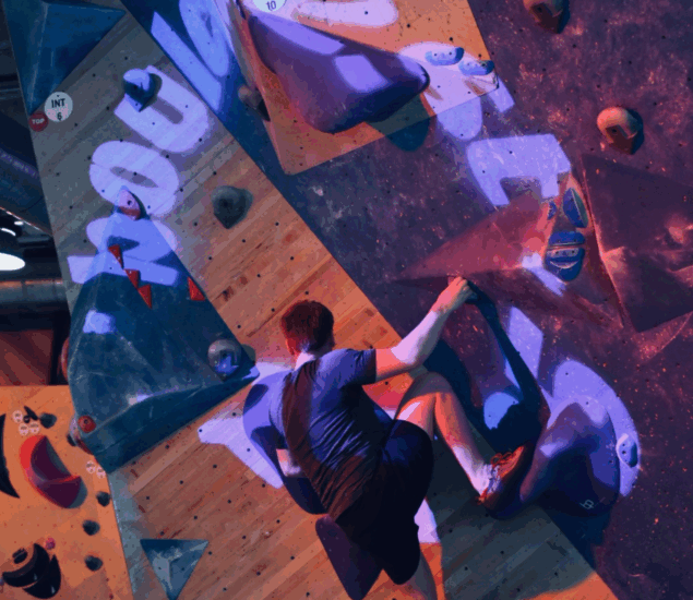 A person wearing a dark shirt and shorts is climbing an indoor bouldering wall at Boulderfest 2025, gripping holds under purple and blue lighting.