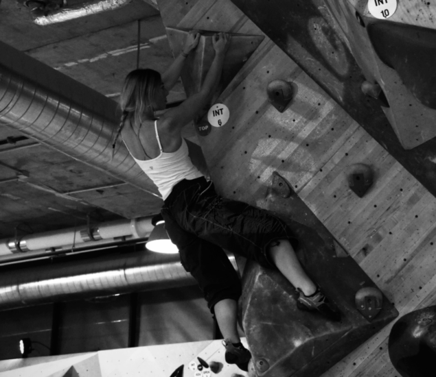 A woman in a tank top and pants climbs an indoor bouldering wall at Boulderfest 2025 in DC, gripping holds with determination. Industrial pipes and ceiling lights form the backdrop in this striking black and white photo.