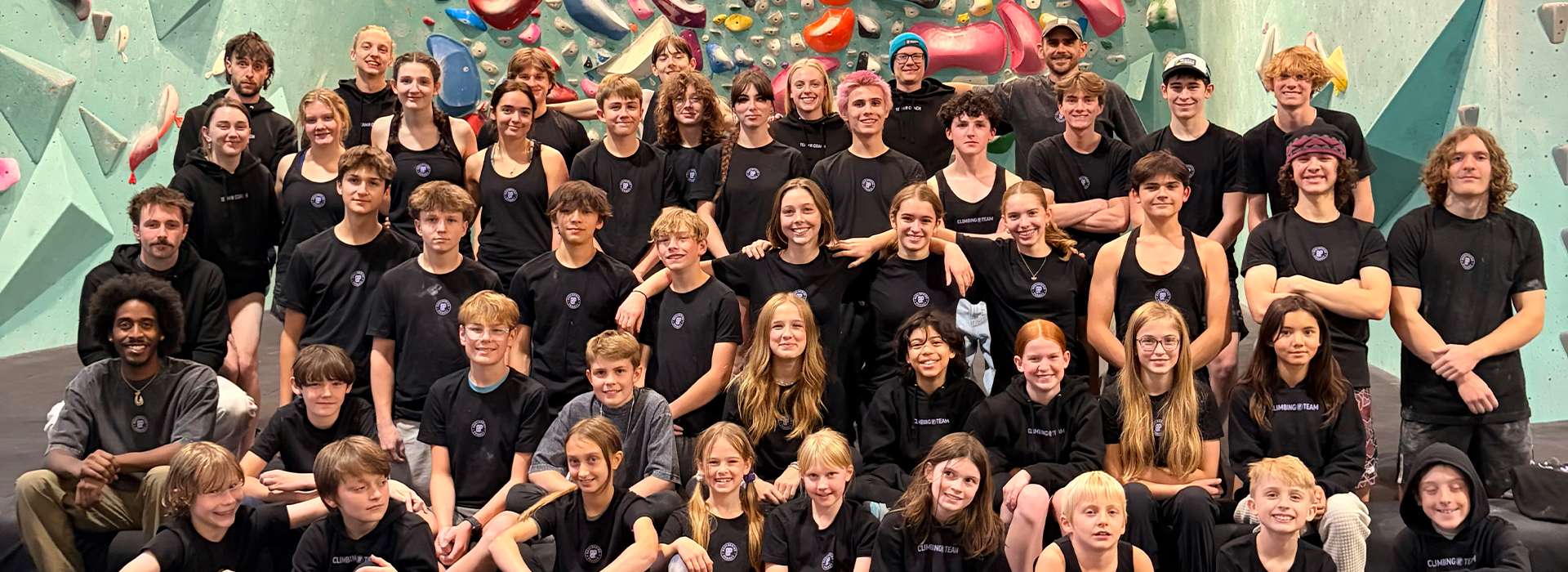 A large group of young people and adults, all wearing matching black shirts, pose and smile together as the Climb Team in front of an indoor climbing wall with colorful holds in Minneapolis.