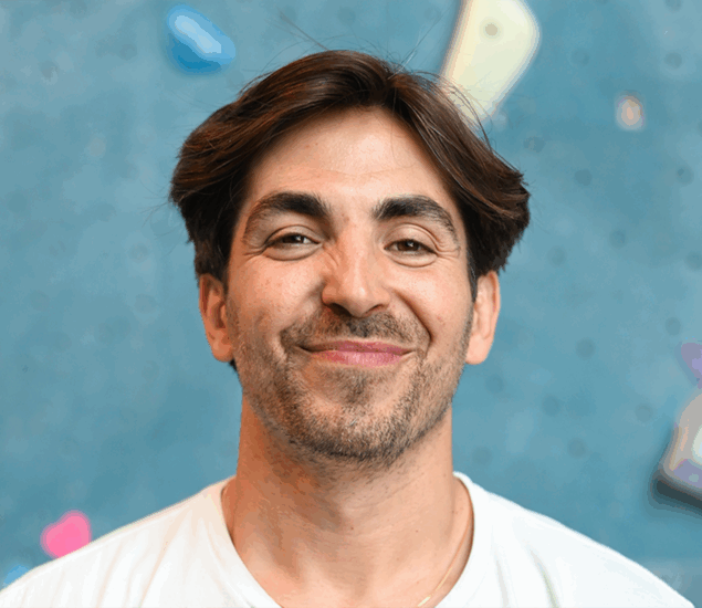 A man with brown hair and light facial hair smiles warmly, wearing a white shirt. The blurred blue background suggests an indoor rock climbing wall—perfect for Personal Climbing Coaching Austin sessions.