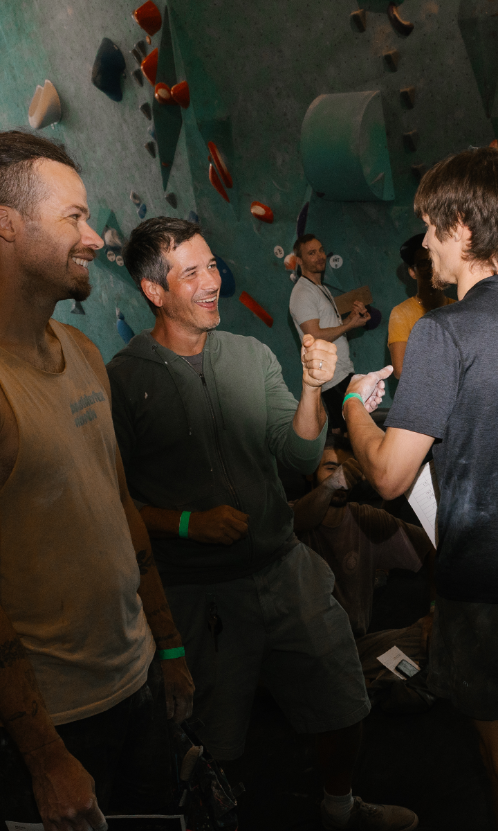 A group of men smiling and giving fist bumps in an indoor climbing gym, with colorful climbing holds on the wall—a perfect moment from the Adult Climbing League.