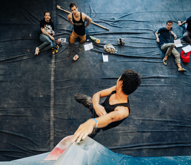 A climber in a black tank top grips a blue climbing wall in a Seattle gym, viewed from above. Three people sit on mats below, watching and surrounded by climbing gear and papers—perfect for personal coaching sessions.
