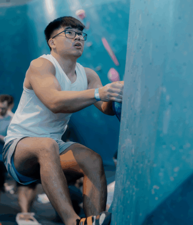 A young man wearing glasses, a white tank top, and shorts is climbing an indoor rock wall, gripping a blue hold. Other climbers and people are blurred in the background, highlighting the focus seen in personal coaching.