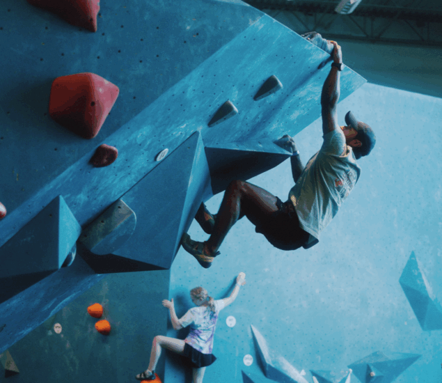 Two people are bouldering indoors on blue climbing walls with red and orange holds. One climber in the foreground hangs from an overhang, benefiting from personal coaching, while another climbs in the background. Both wear athletic clothing.