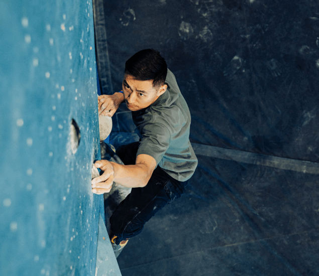 A man climbs an indoor rock wall, gripping holds with both hands and looking up with focus. The background shows padded flooring and chalk marks, reflecting the benefits of personal coaching in all markets.