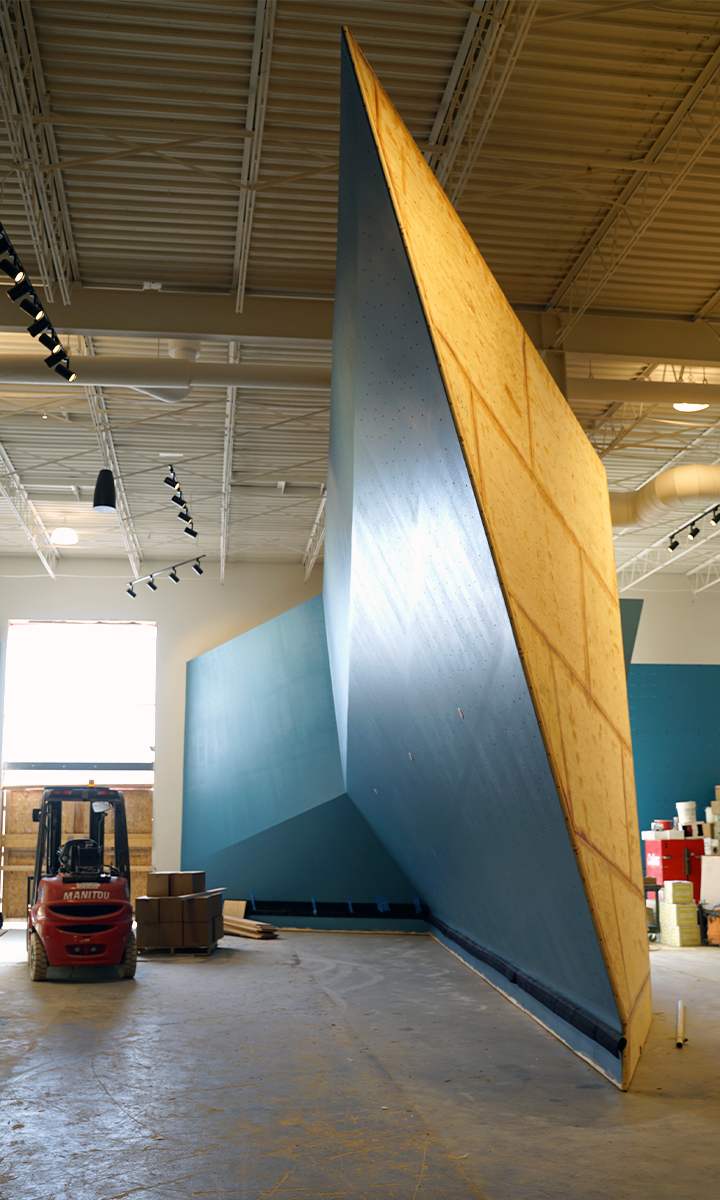 A large, blue indoor climbing wall is being installed in a spacious St Paul rock climbing gym, with a forklift parked nearby and scattered boxes on the floor. Natural light streams in through windows, illuminating the scene.