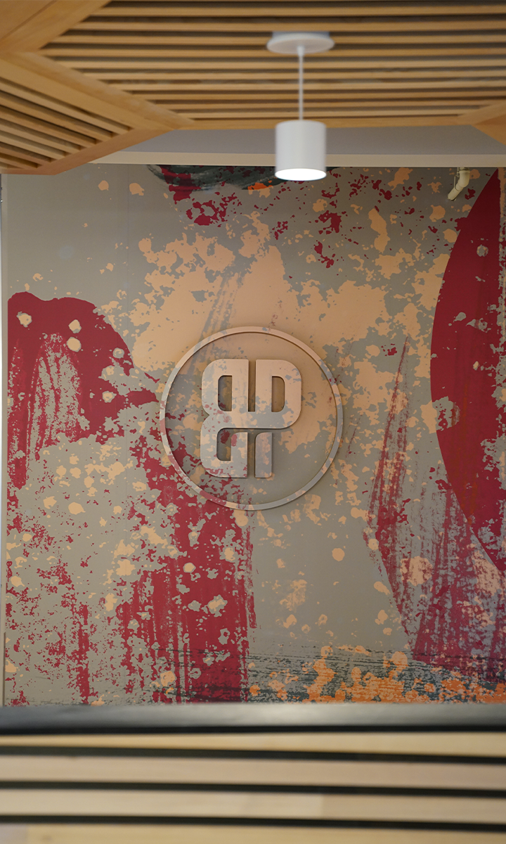 An abstract wall mural with splashes of red, peach, and gray paint, featuring a raised circular emblem with interlocking geometric shapes—perfect for inspiring climbers at a St Paul rock climbing gym. A white ceiling light and wooden slatted panels are above.