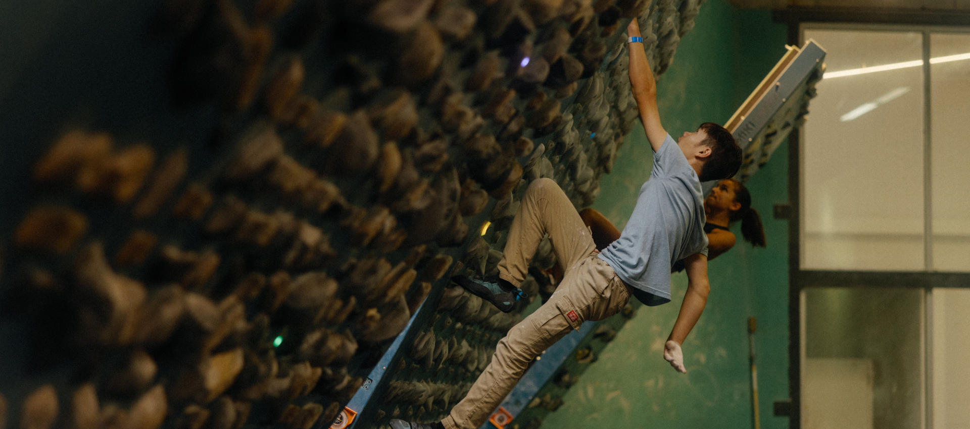 Two people are climbing on an indoor bouldering wall equipped with various handholds and a climbing training board. Both wear climbing gear and focus on ascending, while the greenish wall and large window are visible in the background.