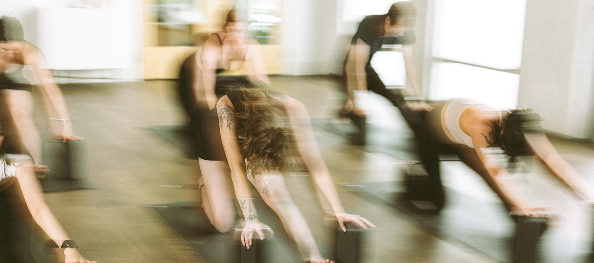 A group of people in blurred motion practicing yoga indoors, each kneeling on mats and reaching forward with their hands on yoga blocks, enhancing flexibility and mental clarity.