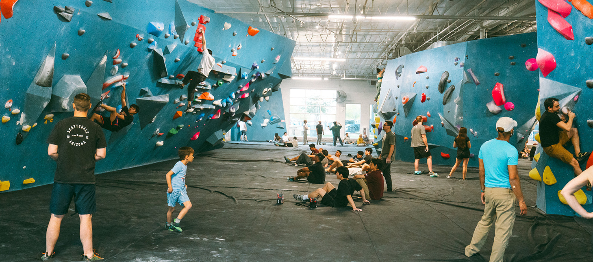 People of various ages climb and gather in a large indoor bouldering gym with blue bouldering walls, colorful holds, and padded flooring. Some are climbing different types of bouldering walls, while others watch, talk, or rest on the mats.