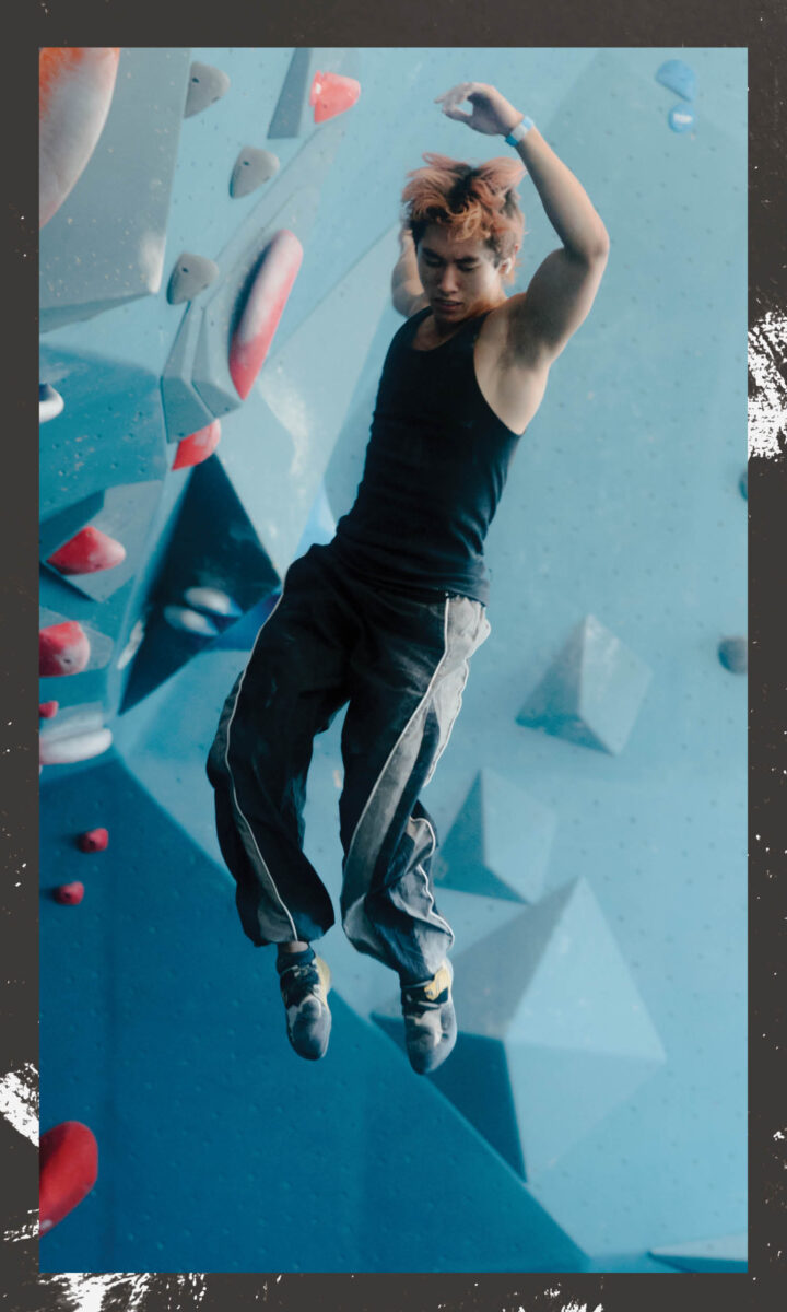 A person in a black tank top and gray pants is captured mid-air in an indoor climbing gym, surrounded by geometric climbing holds on a blue wall—an exciting scene to experience with a fall membership.