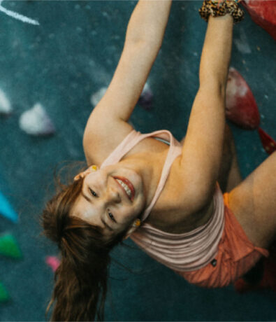 A young woman with a ponytail and wearing a tank top and shorts smiles up at the camera while climbing an indoor rock wall with colorful holds, capturing the fun spirit of a summer camp adventure.