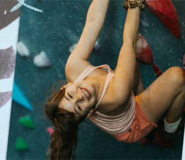 A young woman with a ponytail and wearing a tank top and shorts smiles up at the camera while climbing an indoor rock wall with colorful holds, capturing the fun spirit of a summer camp adventure.