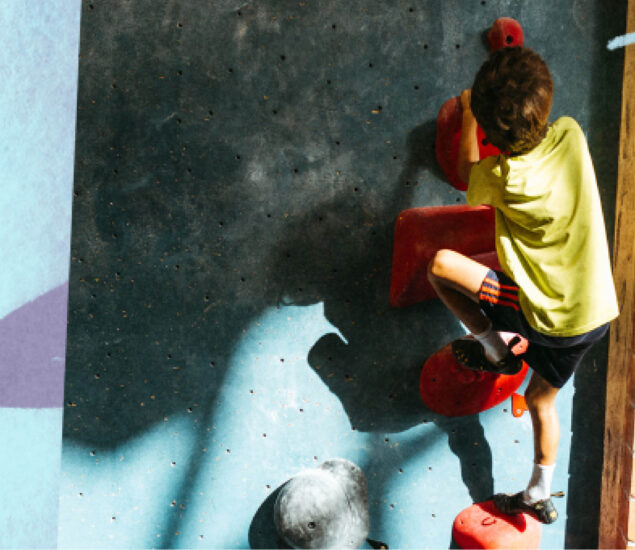 At summer camp, a child in a yellow shirt and striped socks climbs a blue and red indoor rock climbing wall, gripping holds with both hands and feet as sunlight casts playful shadows on the wall.