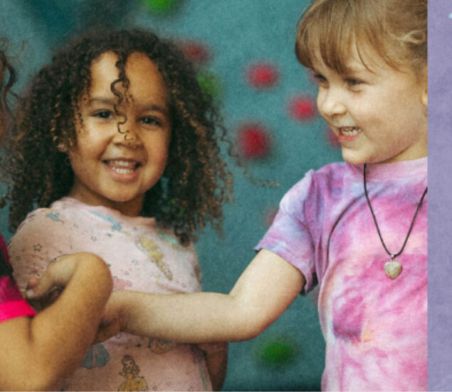 Two young children smile and hold hands at summer camp. One has curly hair and a light pink shirt with cartoon characters; the other sports straight hair, a tie-dye shirt, and a heart-shaped necklace. A blurred, colorful background is visible.