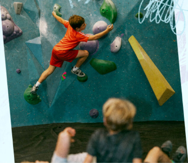 A young boy in a red outfit climbs a colorful indoor bouldering wall at summer camp, while two children watch from below on a padded floor. The wall features green, purple, and yellow climbing holds.