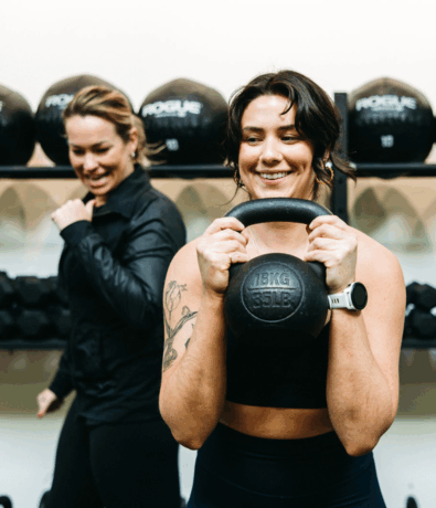 Two women are at a gym. One in front, smiling, holds a 35 lb kettlebell with both hands. Another woman smiles in the background. Both appear to be enjoying their fall membership workout session.