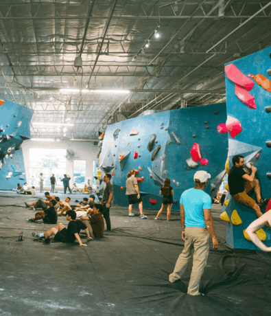 People are bouldering and socializing in a busy indoor climbing gym with colorful bouldering walls, including different types of bouldering walls on blue walls and mats covering the floor. Some climbers learn how to use bouldering walls while others watch or rest.