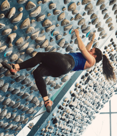 A woman in a blue tank top and black leggings climbs an indoor bouldering wall covered with numerous handholds and footholds, similar to those found on a climbing training board. She reaches up with her right hand, her ponytail swinging.