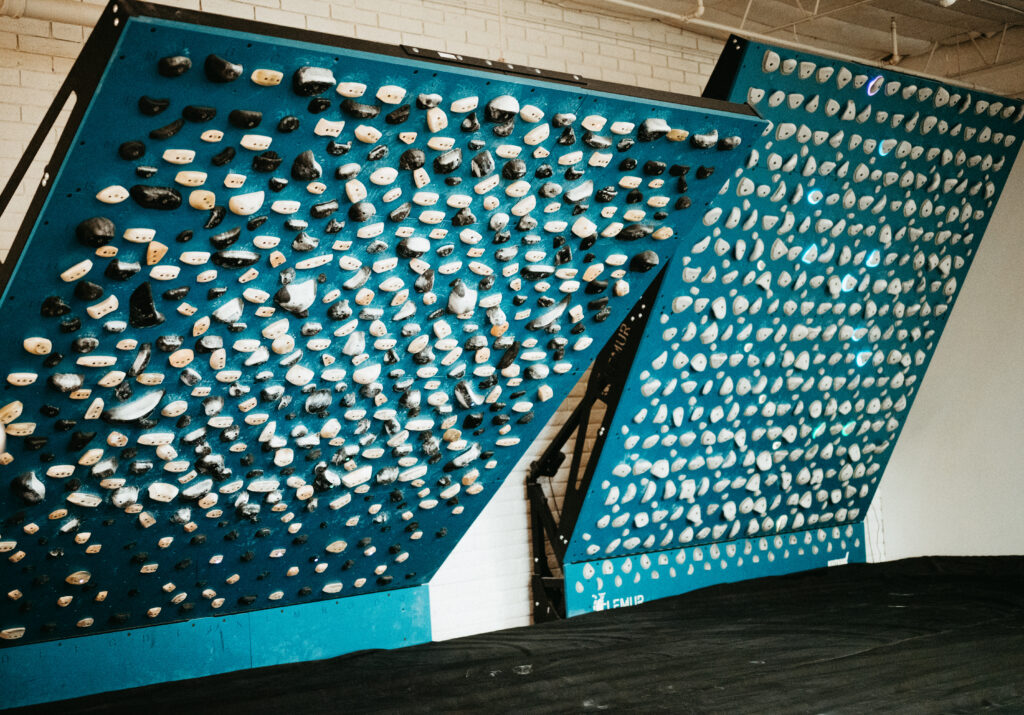 Two blue climbing training boards with numerous white and black holds arranged in grids, set at steep angles over a padded floor in a gym with white brick walls and a visible ceiling.