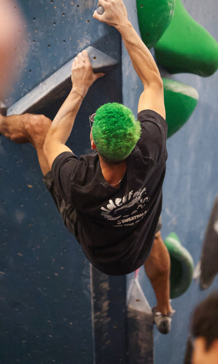 A person with bright green hair climbs an indoor bouldering wall at Boulderfest 2025 in Boston, gripping holds with both hands and wearing a black t-shirt and shorts. Green climbing holds are visible on the wall.