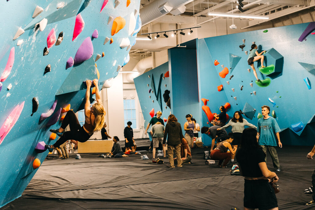 People climb colorful bouldering walls while others watch or prepare. The gym is busy with climbers of various ages exploring different types of bouldering walls and engaging with large, blue climbing structures under bright lighting.