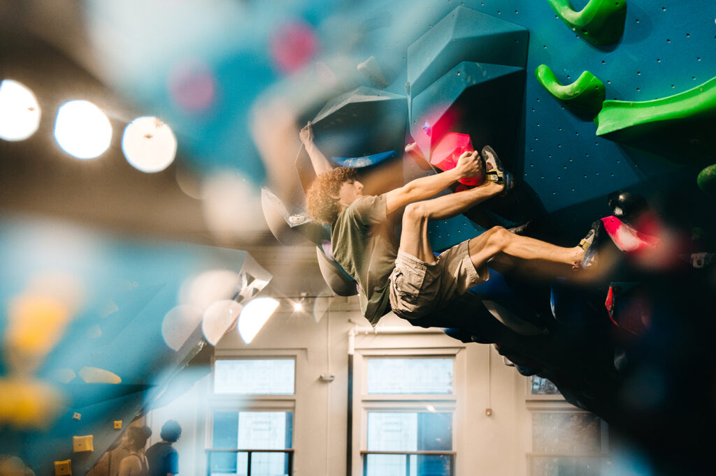 A person with curly hair and shorts climbs vibrant bouldering walls, highlighting the unique features and types of holds. The image is dynamic, showing focus and movement, with a blurred foreground and bright lighting in the background.