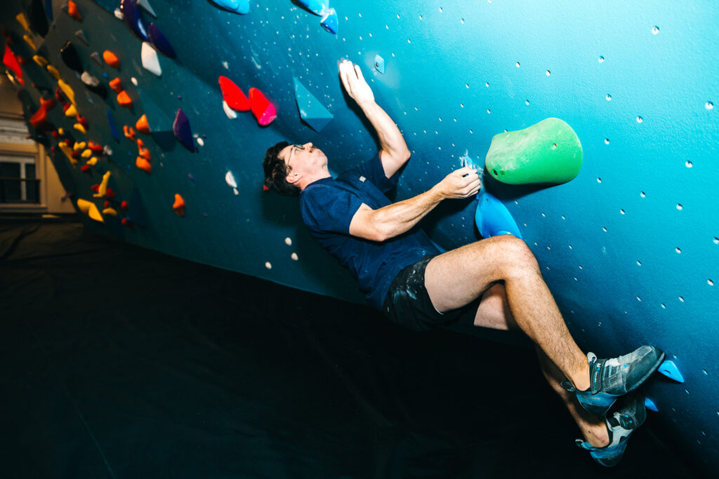 A man wearing a navy shirt and shorts is indoor bouldering on a climbing wall, gripping colorful holds and leaning back with his feet on the wall. A climbing training board is mounted nearby, and the padded floor below ensures safety.