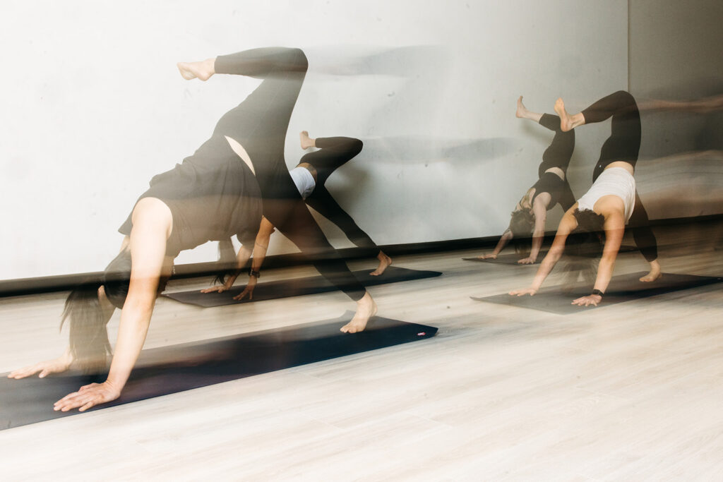 Three people practice heated yoga indoors, each on a mat performing a variation of the three-legged downward dog pose. Motion blur adds a dynamic effect, highlighting flexibility. The room features light wood floors and white walls.