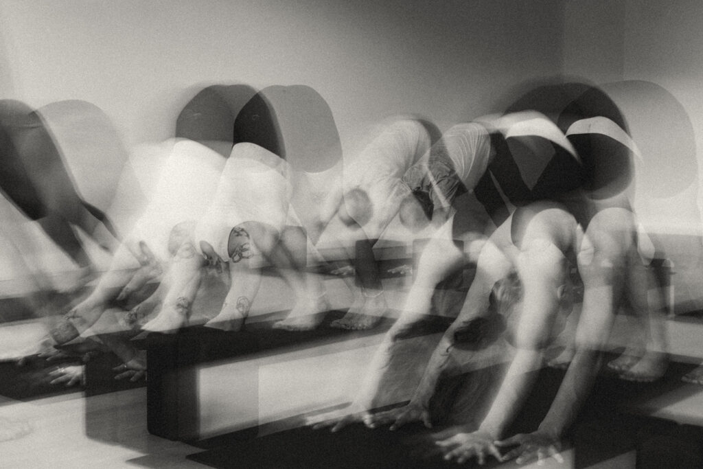A group of people in a Hot Fusion Yoga class perform the downward dog pose on mats. The black and white image uses motion blur, creating a layered, dynamic effect as bodies overlap in movement.