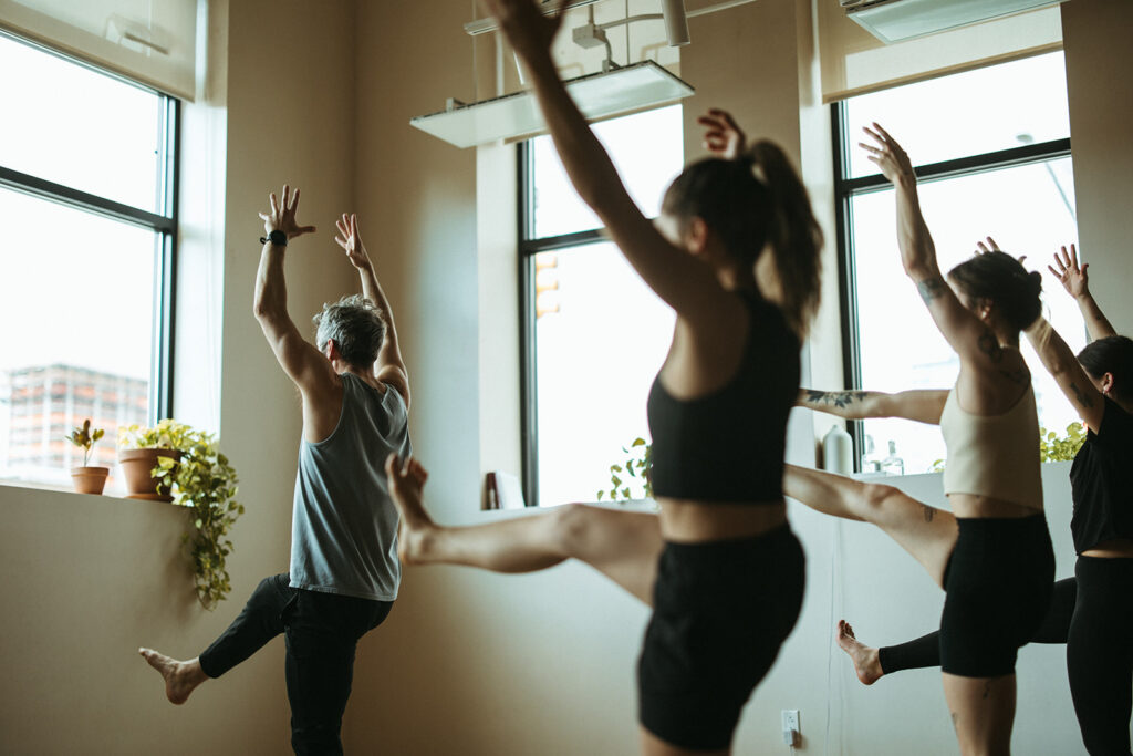 A group of people in athletic wear practice Hot Fusion Yoga, raising one leg and both arms, in a bright room with large windows and potted plants on the windowsill at Bouldering Project.